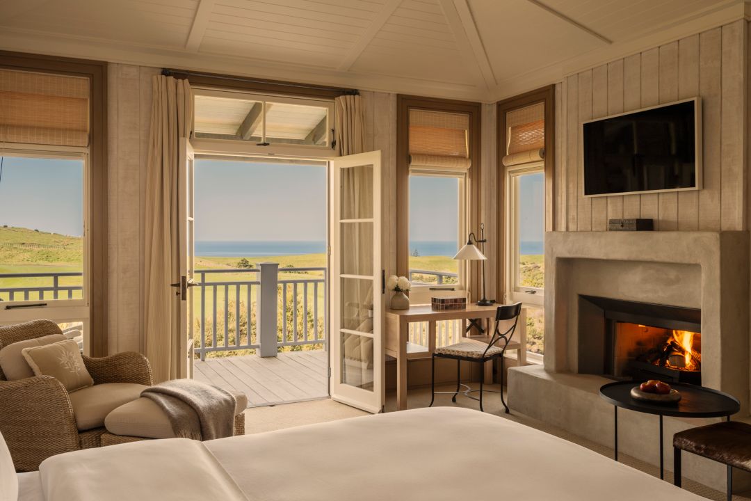 A hotel suite with fireplace, plush bed, armchair, desk and doors opening out to a private balcony with golf course and ocean views.  